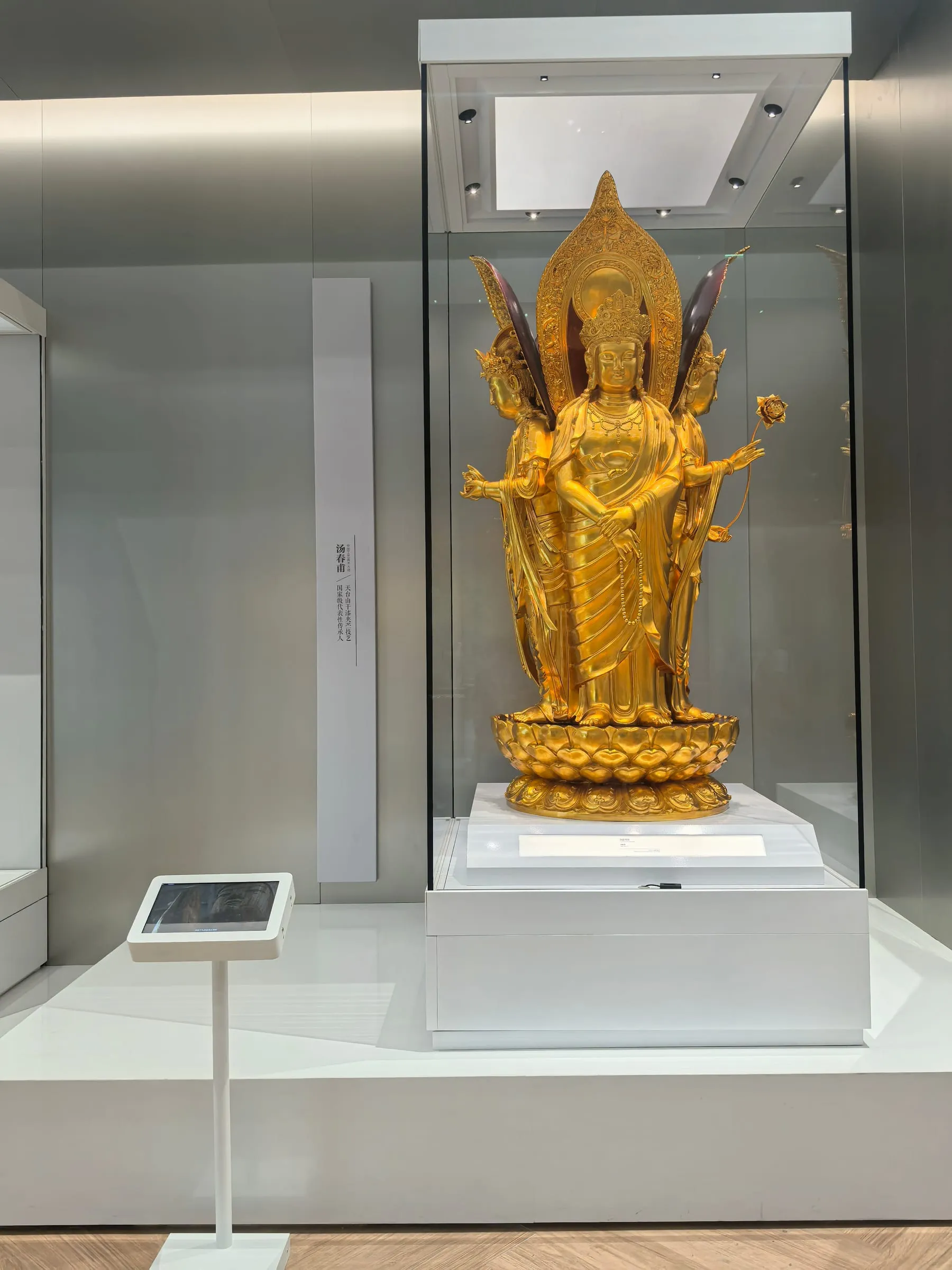 A gilded four-faced Guanyin standing in a glass case in a museum gallery.